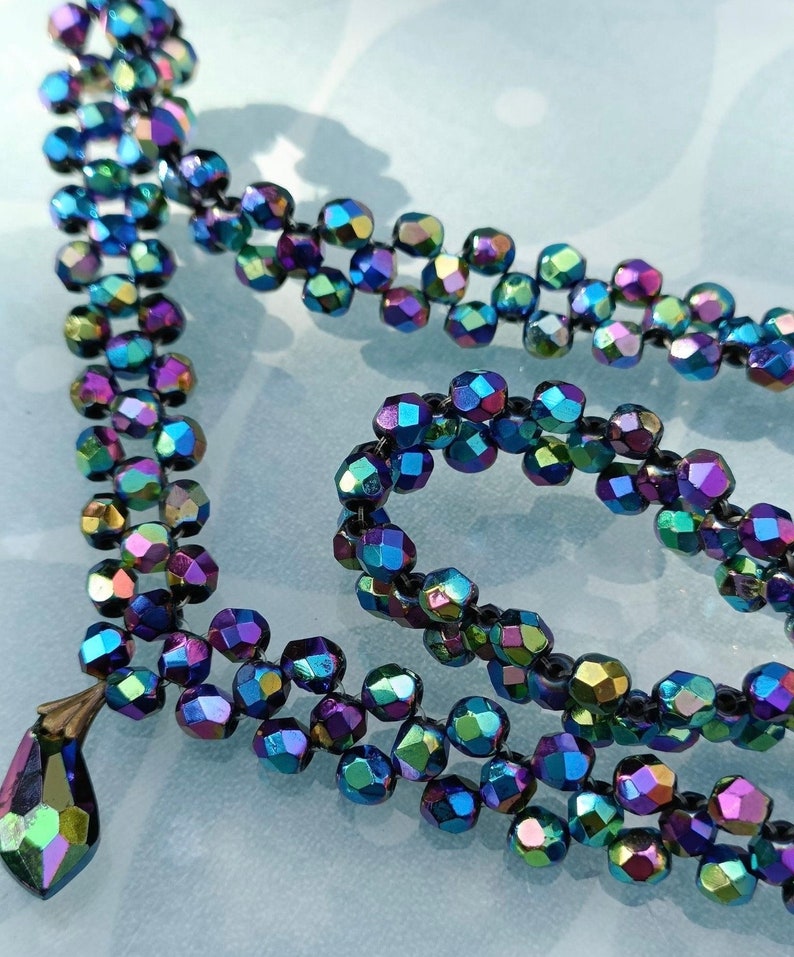 May include: A multi-strand necklace made of iridescent, faceted glass beads. The beads are dark blue, purple, and green, and they are strung on a black thread. The necklace has a teardrop-shaped pendant at the end.