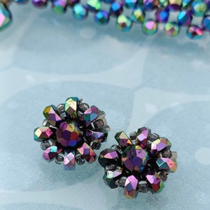 May include: A pair of iridescent earrings with a flower design. The earrings are made of small, faceted beads that are arranged in a circular pattern. The beads are a mix of purple, blue, and green.