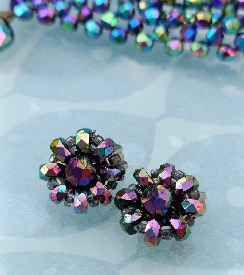 May include: A pair of iridescent earrings with a flower design. The earrings are made of small, faceted beads that are arranged in a circular pattern. The beads are a mix of purple, blue, and green.