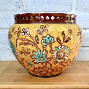 Puede incluir: Un macetero de cerámica amarillo con un borde marrón y un diseño floral intrincado en tonos de marrón, azul y blanco. El macetero tiene una superficie texturizada y un borde festoneado.