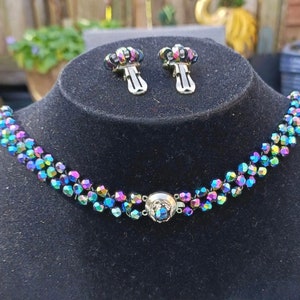 May include: A multi-strand iridescent choker necklace with a matching pair of clip-on earrings. The necklace is made of small, faceted beads that are strung together in a repeating pattern. The earrings are also made of faceted beads and have a simple, elegant design.