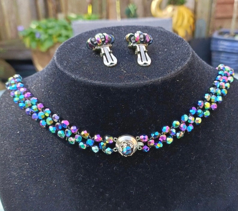 May include: A multi-strand iridescent choker necklace with a matching pair of clip-on earrings. The necklace is made of small, faceted beads that are strung together in a repeating pattern. The earrings are also made of faceted beads and have a simple, elegant design.