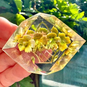 Pode incluir: Broche transparente em forma de diamante com um design floral. O broche apresenta um cesto de flores amarelas e verdes envolto em material transparente. As bordas do broche são douradas e o fundo é preto.