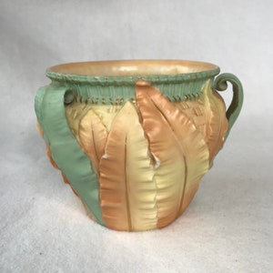May include: A ceramic planter with two handles, decorated with a raised design of green and peach leaves. The planter has a gold trim around the top.
