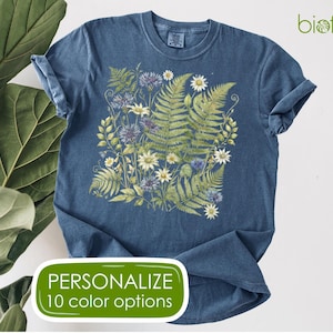 May include: A blue t-shirt featuring a botanical design of ferns, daisies, and other wildflowers. The shirt has rolled-up sleeves and the text "PERSONALIZE 10 color options" in a green oval. The brand name "biofillia" is in the upper right corner.
