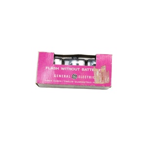 May include: A vintage General Electric flash cube box. The pink box reads "FLASH WITHOUT BATTERIES" and contains three flash cubes. The box has some wear and tear, with the General Electric logo visible.