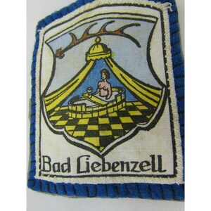 May include: A square patch with a blue border and a white center. The center features a crest with a woman in a bath, a yellow canopy, and the text "Bad Liebenzell". The background is a checkered pattern.
