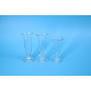 May include: Three clear glass graduated cylinders of different sizes, each with a flared top and a base. The cylinders have measurement markings in millilitres. The background is a solid light blue.