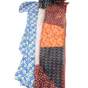May include: A colorful scarf with a patchwork design. The scarf features sections of white and blue geometric patterns, floral prints in orange and white, and a dark red floral pattern. The scarf is made of a lightweight fabric.