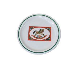 May include: White ceramic plate with a green rim. The center of the plate features a red rectangle with a green border. Inside the rectangle is a colorful illustration of a rocking horse with a green, red, and white design. The text "Christmas Collection" is printed in blue at the bottom of the rectangle.