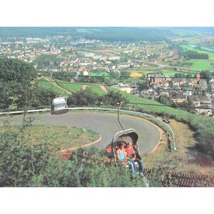 May include: A vintage postcard depicting a scenic view of a town from a mountaintop. Two cable cars are visible, one with three people riding in it.
