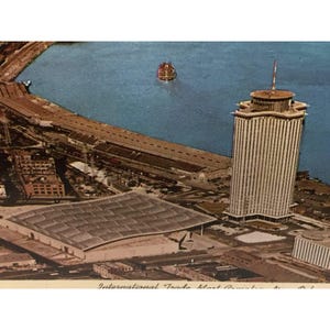 Könnte beinhalten: Eine Luftaufnahme des International Trade Mart in New Orleans, Louisiana. Das Gebäude ist ein großes, modernes Bauwerk mit einem Flachdach und einem markanten Design. Im Hintergrund ist ein Fluss zu sehen, auf dem ein Boot fährt.