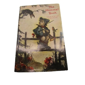Può includere: La copertina di un libro intitolato "The Hummel Book" con un'illustrazione di un giovane ragazzo seduto su una recinzione con un uccello e un corvo. Il testo sotto il titolo recita "From the life and work of Berta Hummel."