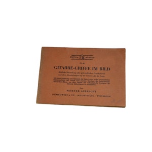 Può includere: Un libro marrone con il titolo "GITARRE-GRIFFE IM BILD" e il testo "EDITION WACHTLER Nr. 46" sulla copertina. Il libro è per l'insegnamento della chitarra ed è pubblicato da DOMKOWSKY & CO. MUSIKVERLAG WIESBADEN.