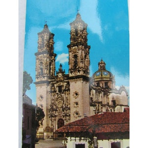 May include: A vintage postcard featuring a detailed view of a large, ornate church with two tall towers and a dome. The church is made of light-colored stone and has intricate carvings and details. The sky is a bright blue.