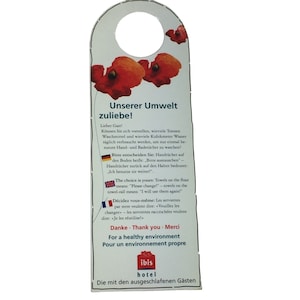 May include: A white door hanger with a red poppy flower design. The text on the hanger reads "Unserer Umwelt zuliebe!" which translates to "For the sake of our environment!" in English. The text also includes information about towel usage and a message in German, English, and French encouraging guests to be mindful of their environmental impact. The hanger also features the ibis hotel logo.