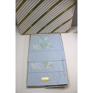 May include: A set of two blue pillowcases with white embroidery. The top pillowcase says "His" and the bottom pillowcase says "Hers". Both pillowcases have a small floral design with green leaves and white flowers. The pillowcases are in a white box with a green and white striped pattern.