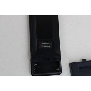 May include: Black remote control with a battery compartment open, revealing two spring-loaded battery contacts. The remote has the text "VCR964N PART NO. 076G041060 MADE IN SINGAPORE" printed on it.