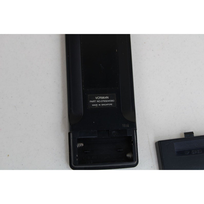 May include: Black remote control with a battery compartment open, revealing two spring-loaded battery contacts. The remote has the text "VCR964N PART NO. 076G041060 MADE IN SINGAPORE" printed on it.