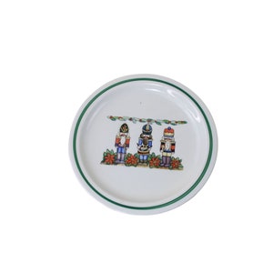 May include: A white ceramic plate with a green rim features a festive design of three nutcracker figurines in colorful attire. The nutcrackers are standing in front of a garland of holly and poinsettia flowers.