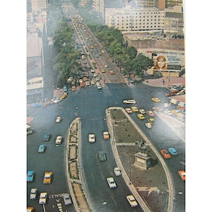 May include: An aerial view of a busy city intersection with cars driving in all directions. The intersection is surrounded by buildings and trees. There is a statue in the middle of the intersection.