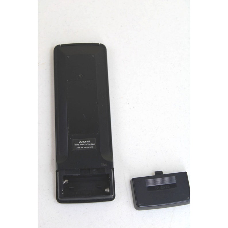 May include: Black remote control with a battery compartment open, revealing two AA batteries. The remote has the text "VCR964N PART NO. L7050304R01 MADE IN SINGAPORE" printed on it. The battery cover is separate and black.