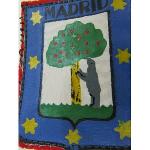 May include: A blue and white patch featuring the word "MADRID" at the top. The patch has a shield design with a green tree with red apples, a yellow trunk, and a gray bear. Yellow stars are on the blue background.