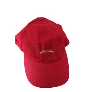 Puede incluir: Gorra de béisbol roja con la palabra "Paddling" bordada en hilo rojo más oscuro. Debajo, un diseño dorado de remos cruzados. La gorra tiene una visera curva y una corona redondeada.