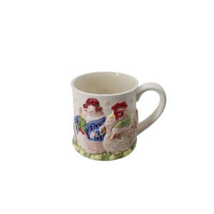 Könnte beinhalten: Eine cremefarbene Keramiktasse mit weißem Henkel. Die Tasse hat ein 3D-Reliefdesign eines Schweins und eines Huhns, die beide Kleidung tragen. Das Schwein trägt eine blaue Jacke und einen roten Hut, während das Huhn einen grünen Schal hat.