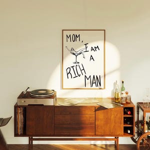 May include: A framed print with a black and white illustration of a woman reclining in a martini glass. The text "MOM, I AM A RICH MAN" is written in black below the illustration.