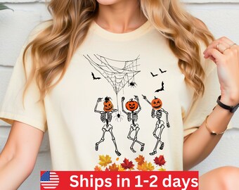 May include: Cream-coloured t-shirt with a Halloween design. The graphic features three dancing skeletons with pumpkin heads, a spider web, bats, and spiders. Autumn leaves are at the bottom of the design.