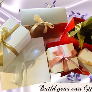 Op de afbeelding: Een verzameling cadeaudozen in verschillende maten en kleuren, waaronder wit, goud, rood en roze. De dozen zijn gebonden met linten en strikken, en sommige bevatten tissuepapier. De tekst "Build your own Gift Box" is zichtbaar onderaan de afbeelding.