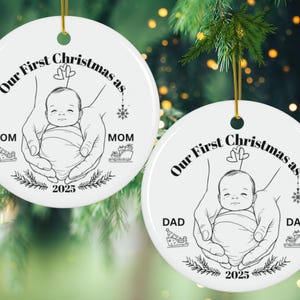 May include: Two white ceramic Christmas ornaments with gold hanging strings. Each ornament features a black and white illustration of a baby held in hands, with text that reads "Our First Christmas as" and "2025". One says "Mom" and the other says "Dad".