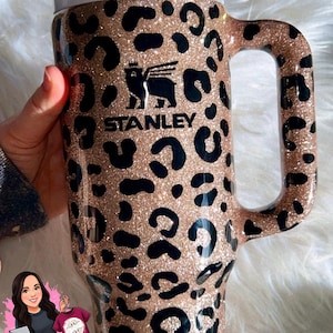 May include: A large, insulated tumbler with a handle, featuring a leopard print design in black and gold glitter. The Stanley logo is visible. Includes a white lid and straw. Ideal for cold beverages.