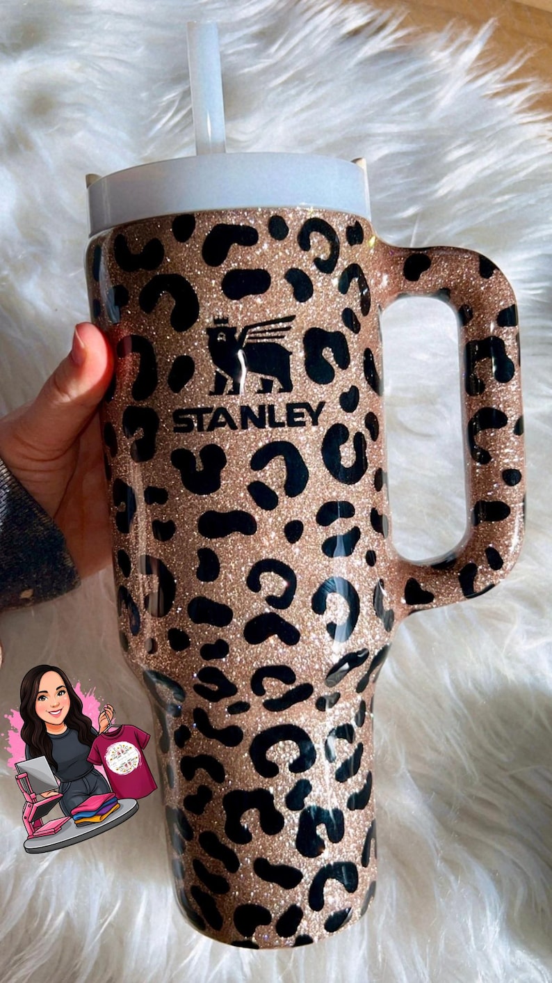May include: A large, insulated tumbler with a handle, featuring a leopard print design in black and gold glitter. The Stanley logo is visible. Includes a white lid and straw. Ideal for cold beverages.