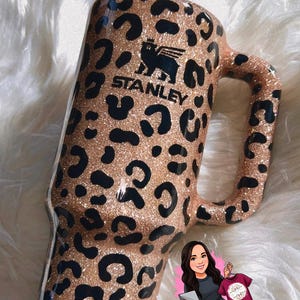 May include: A glittery, leopard-print Stanley tumbler with a handle and a white lid. The tumbler features the Stanley logo in black. The design includes black spots on a tan background. A white straw is inserted into the lid.