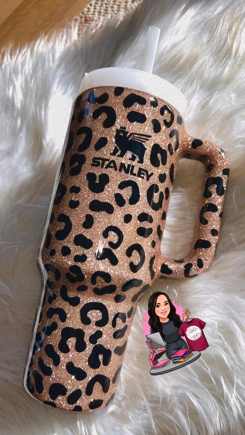 May include: A glittery, leopard-print Stanley tumbler with a handle and a white lid. The tumbler features the Stanley logo in black. The design includes black spots on a tan background. A white straw is inserted into the lid.