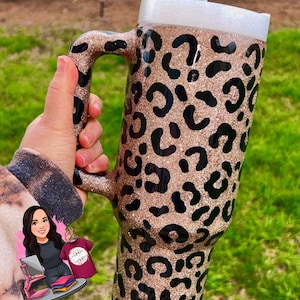 May include: A large, glittery tumbler with a handle, featuring a leopard print design in black and gold. The tumbler has a white lid and a clear straw. The background is blurred green grass.