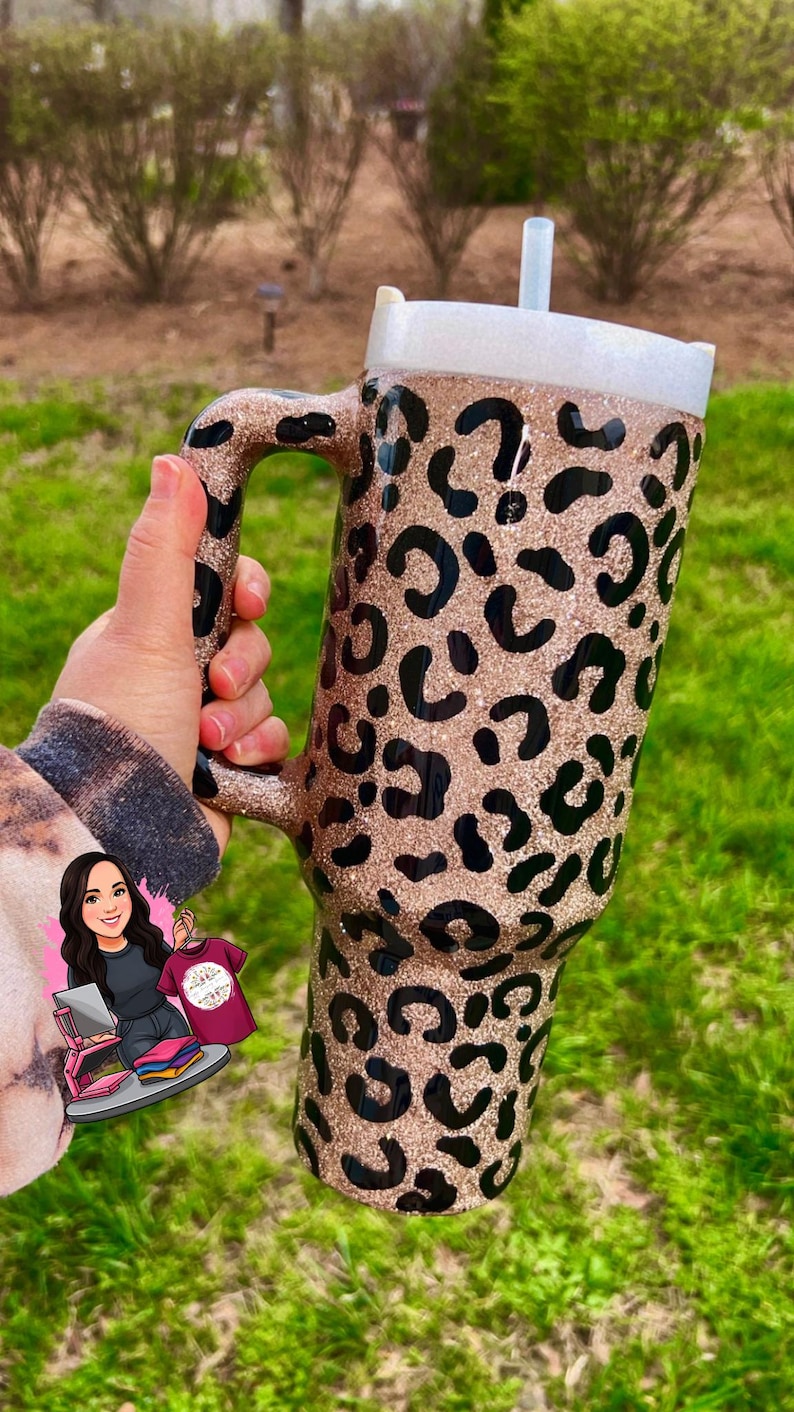 May include: A large, glittery tumbler with a handle, featuring a leopard print design in black and gold. The tumbler has a white lid and a clear straw. The background is blurred green grass.