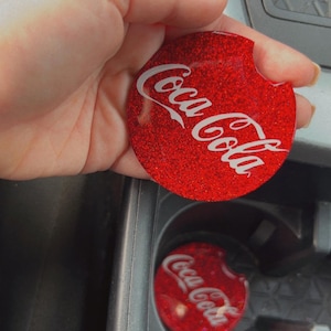 May include: Red glitter car coaster with the Coca-Cola logo in white. The coaster is round and fits in a car's cup holder. A second coaster is visible in the car's cup holder.