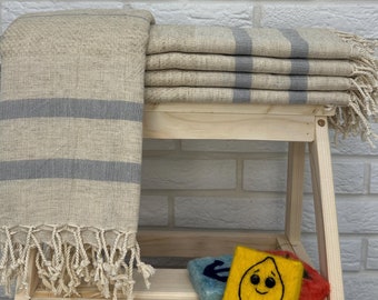 Puede incluir: Una pila de toallas rayadas beige y grises con flecos, colocadas en un estante de madera. Las toallas están dobladas cuidadosamente y tienen una textura sutil.