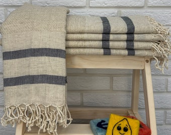 Puede incluir: Una pila de toallas rayadas beige y negras con flecos en un estante de madera. Las toallas están dobladas y apiladas unas sobre otras. Hay tres pequeñas decoraciones de fieltro en el estante debajo de las toallas.