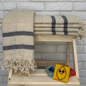 May include: A stack of beige and black striped towels with fringe on a wooden shelf. The towels are folded and stacked on top of each other. There is a small yellow felt lemon with a smiley face on it in the foreground.