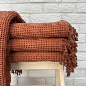 May include: A stack of four waffle weave towels in a terracotta orange color with fringe trim. The towels are folded and stacked on a wooden stool.