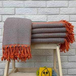 Op de afbeelding: Een stapel van zes oranje en grijze handdoeken met oranje franjes. De handdoeken zijn gevouwen en gestapeld op een houten plank.