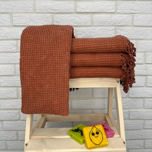 May include: A stack of four rust-colored waffle weave towels with fringe on the ends. The towels are folded and stacked on a wooden stool.