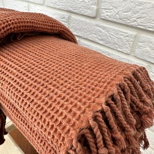 May include: A waffle weave throw blanket in a terracotta color with a fringe border. The blanket is folded and draped over a white brick wall.