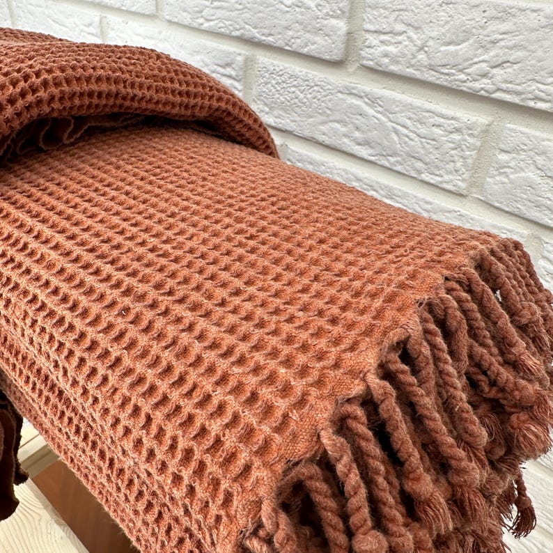 May include: A waffle weave throw blanket in a terracotta color with a fringe border. The blanket is folded and draped over a white brick wall.