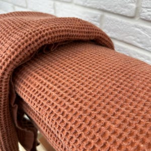 May include: A close-up of a rust-colored waffle weave throw blanket folded on a white brick surface. The blanket is soft and textured.