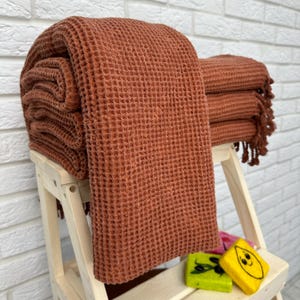 May include: A stack of terracotta waffle weave towels with fringed edges. The towels are folded and displayed on a wooden step stool.